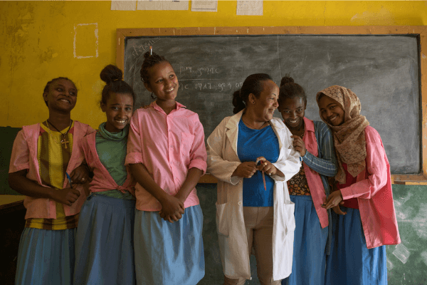 5 school girls laughing and smiling standing with their teacher in a classroom